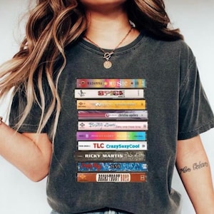 Op de afbeelding: Donkergrijs t-shirt met een print van retro cassettebandjes, met namen van artiesten als Madonna en Spice Girls. Het ontwerp is gedrukt op een casual t-shirt, ideaal voor muziekliefhebbers.