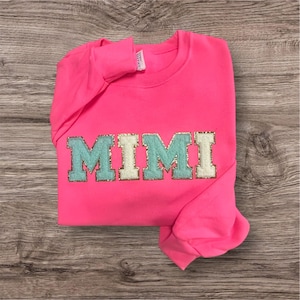 May include: A neon pink long-sleeve sweatshirt featuring the word MIMI in large block letters across the chest. The letters are crafted with a textured, glittery gold border and alternating light blue and cream-colored fabric centers.