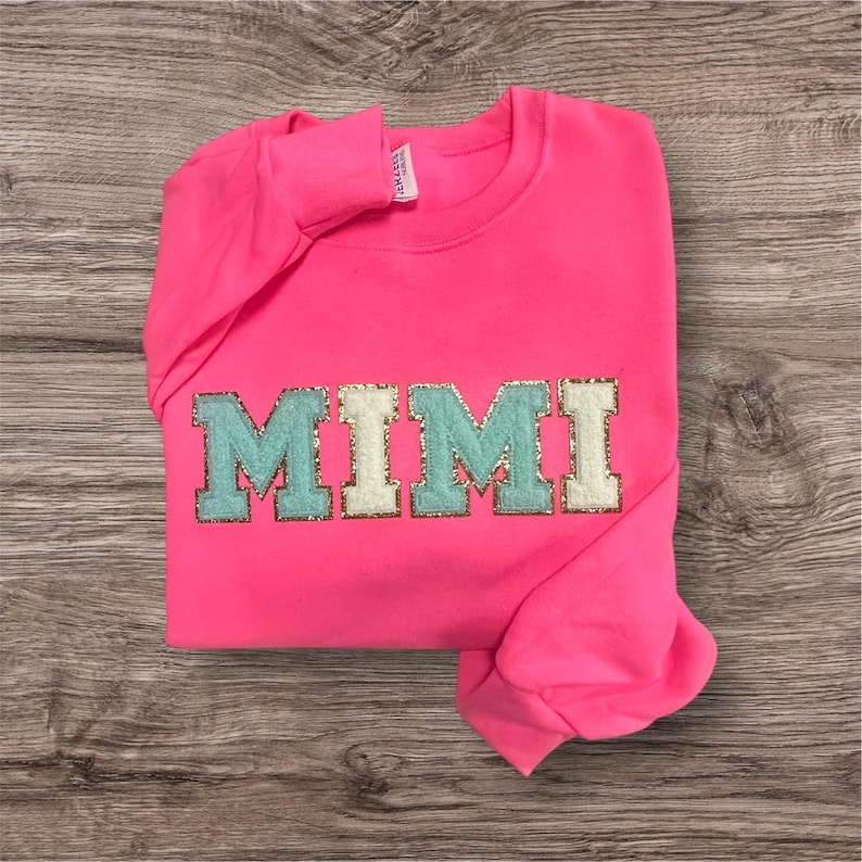 May include: A neon pink long-sleeve sweatshirt featuring the word MIMI in large block letters across the chest. The letters are crafted with a textured, glittery gold border and alternating light blue and cream-colored fabric centers.