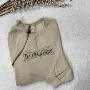 K&ouml;nnte beinhalten: Beiges Sweatshirt mit dem gestickten Wort "Disneyland" in einem farbenfrohen Blockbuchstaben-Design. Das Sweatshirt liegt gefaltet auf einer wei&szlig;en Oberfl&auml;che, mit einem Zweig wei&szlig;er Blumen im Hintergrund.