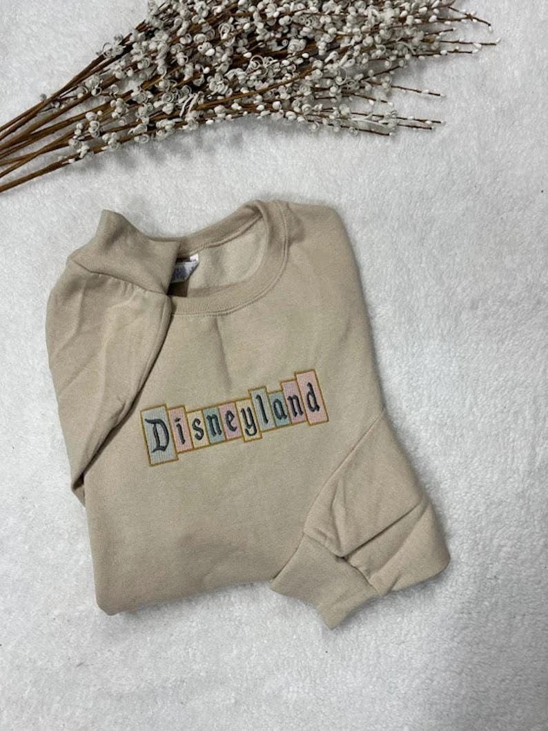K&ouml;nnte beinhalten: Beiges Sweatshirt mit dem gestickten Wort "Disneyland" in einem farbenfrohen Blockbuchstaben-Design. Das Sweatshirt liegt gefaltet auf einer wei&szlig;en Oberfl&auml;che, mit einem Zweig wei&szlig;er Blumen im Hintergrund.