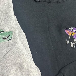 May include: Two sweatshirts, one gray and one black, each embroidered with a cartoon car design. The gray sweatshirt features a green car, while the black one has a purple car with flames.