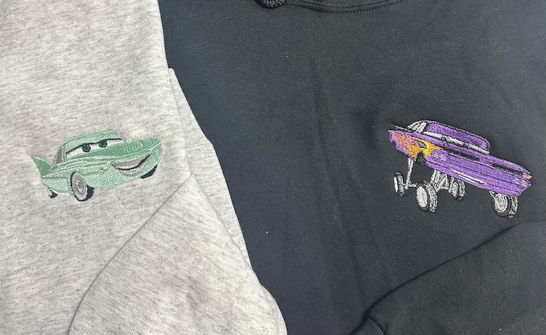 May include: Two sweatshirts, one gray and one black, each embroidered with a cartoon car design. The gray sweatshirt features a green car, while the black one has a purple car with flames.