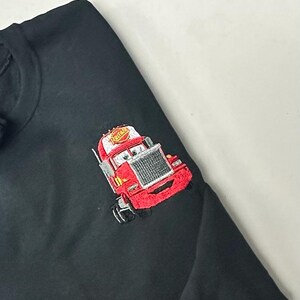 May include: Black sweatshirt featuring an embroidered red cartoon truck with a smiling face and the word "Rusteze" on the cab. The truck has a silver grill and white eyes. The sweatshirt is made of a soft material.