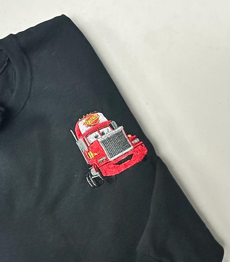 May include: Black sweatshirt featuring an embroidered red cartoon truck with a smiling face and the word "Rusteze" on the cab. The truck has a silver grill and white eyes. The sweatshirt is made of a soft material.