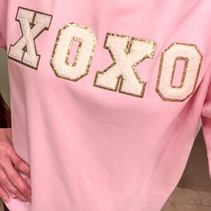 May include: A light pink crewneck sweatshirt featuring the text XOXO across the chest. The letters are rendered in a white, textured material with a gold glitter border, creating a varsity-style appliqu&eacute; effect on the front of the garment.