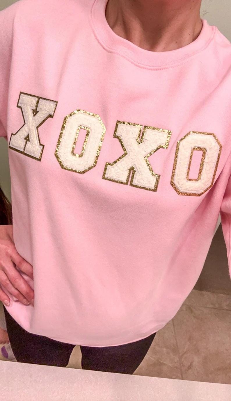 May include: A light pink crewneck sweatshirt featuring the text XOXO across the chest. The letters are rendered in a white, textured material with a gold glitter border, creating a varsity-style appliqu&eacute; effect on the front of the garment.