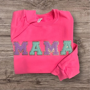 May include: A bright neon pink long-sleeve sweatshirt featuring the word MAMA in large, block-style letters across the chest. The letters alternate between purple and light green, each outlined with a shimmering gold glitter border.