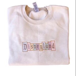 K&ouml;nnte beinhalten: Cremefarbenes Sweatshirt mit dem gestickten Wort "Disneyland" in Pastellfarben auf der Brust. Das Sweatshirt ist ordentlich gefaltet, ein Etikett ist am Kragen sichtbar.