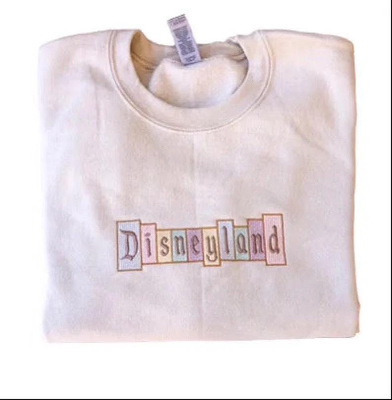 K&ouml;nnte beinhalten: Cremefarbenes Sweatshirt mit dem gestickten Wort "Disneyland" in Pastellfarben auf der Brust. Das Sweatshirt ist ordentlich gefaltet, ein Etikett ist am Kragen sichtbar.