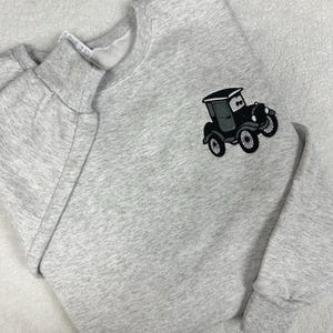 May include: A light gray sweatshirt with a crew neck and long sleeves. An embroidered black and gray vintage car is on the front. The sweatshirt is made of a soft, comfortable material.
