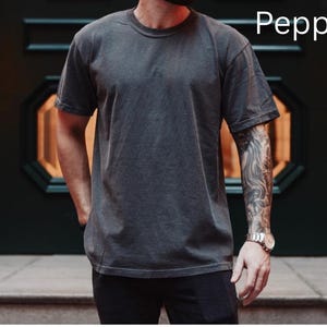 May include: A dark gray, short-sleeved t-shirt with a crew neck. The shirt has a slightly faded appearance and the word "Pepper" is printed in white on the upper right. The shirt is worn with black pants.