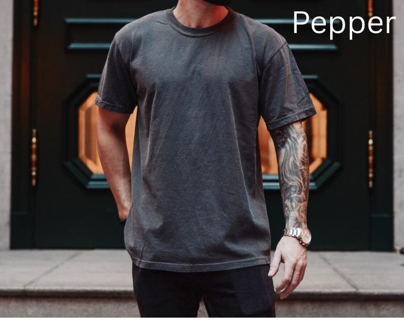 May include: A dark gray, short-sleeved t-shirt with a crew neck. The shirt has a slightly faded appearance and the word "Pepper" is printed in white on the upper right. The shirt is worn with black pants.