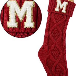 Puede incluir: Un calcetín navideño rojo con la letra "M" blanca con borde dorado. El calcetín está hecho de punto con un diseño de trenzas. La parte superior del calcetín está llena de adornos festivos, incluyendo vegetación y bayas.