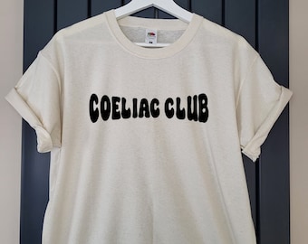 May include: A white t-shirt with the text "COELIAC CLUB" in black, retro-style lettering.