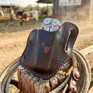 May include: A dark brown leather multi-tool sheath featuring a stamped longhorn skull logo and contrast stitching. The sheath has integrated belt loops and holds a silver metal multi-tool. A coiled rope with a leather and brass buckle is attached.