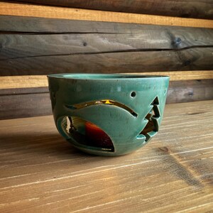 Op de afbeelding: Een teal keramische kandelaar met uitgesneden ontwerpen van een boom en andere vormen. De kandelaar heeft een gouden interieur en staat op een houten oppervlak. Een rode kaars is zichtbaar binnenin.