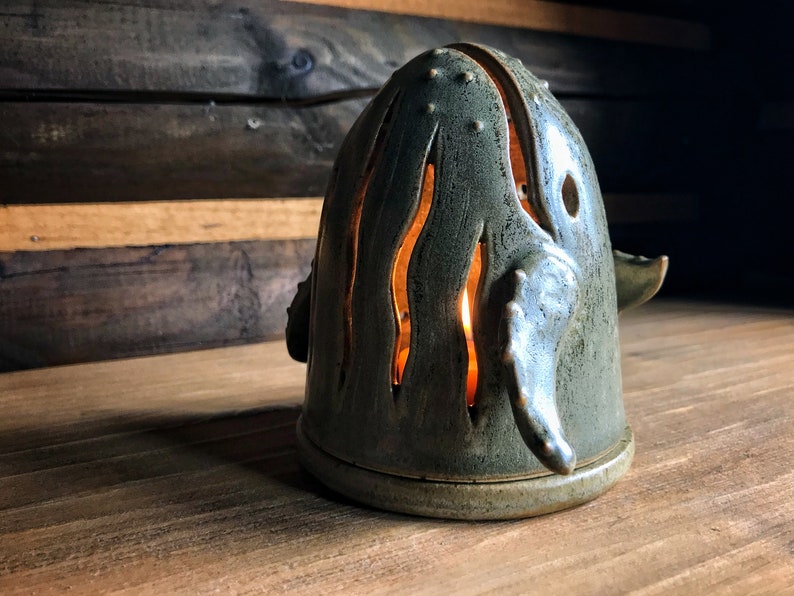 May include: A ceramic whale candle holder with a teal glaze and cut-out details. The candle holder is lit and casting a warm glow.