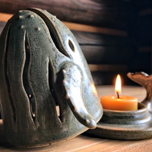 May include: A ceramic whale candle holder with a teal green glaze. The whale is open on the top and has a tail fin on the bottom. A small teal green base holds a lit tea light candle.