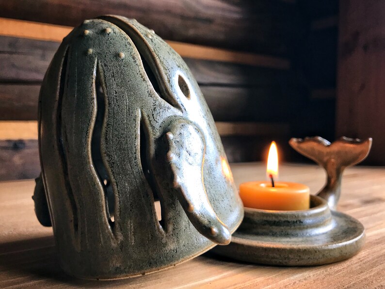 May include: A ceramic whale candle holder with a teal green glaze. The whale is open on the top and has a tail fin on the bottom. A small teal green base holds a lit tea light candle.
