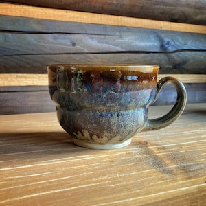 Puede incluir: Una taza de cerámica hecha a mano con un cuerpo redondeado y un asa curva. La taza presenta un esmalte brillante con tonos marrones, azules y verdes. La taza está sobre una superficie de madera.