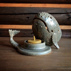 May include: A gray ceramic whale candle holder with a teal glaze. The whale is holding a small, round, yellow candle in its mouth.
