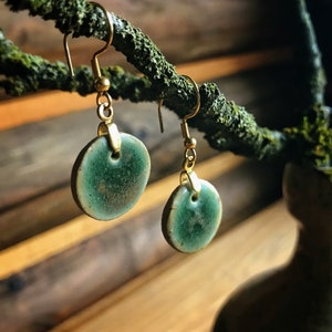 May include: A pair of handmade earrings with a circular, teal-colored ceramic pendant. The earrings have gold-colored hooks and accents. The earrings are hanging from a moss-covered branch, with a wooden background.