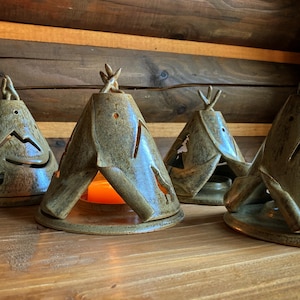 May include: Four ceramic teepee-shaped candle holders in a rustic style. Each holder is a muted green-blue with cut-out designs of mountains and animals. The candle holders are arranged on a wooden surface.