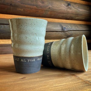 Könnte beinhalten: Zwei handgefertigte Keramikbecher. Einer steht aufrecht, der andere liegt auf der Seite. Die Becher sind hellbeige mit dunklen Sprenkeln und einem dunkelgrauen Boden. Auf den Bechern steht "CAST ALL YOUR ANXIETY" und "WHO GIVES ME STRENGTH".