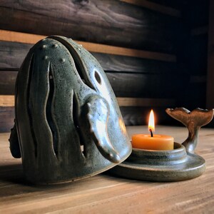 May include: A ceramic whale candle holder with a teal glaze and a cutout design. The whale is open and a small, round, yellow candle is inside. The whale's tail is separate and sits on a base.