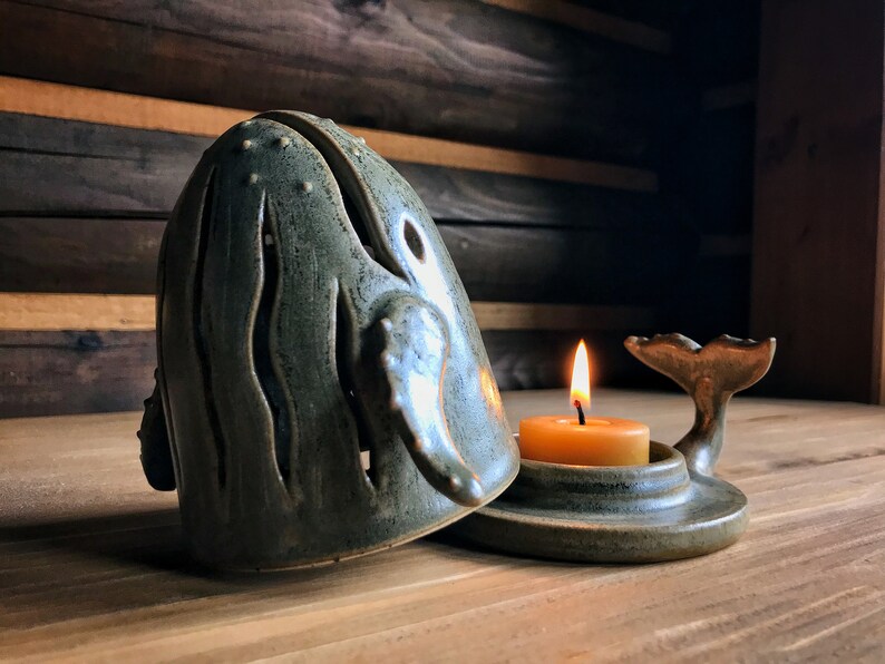 May include: A ceramic whale candle holder with a teal glaze and a cutout design. The whale is open and a small, round, yellow candle is inside. The whale's tail is separate and sits on a base.