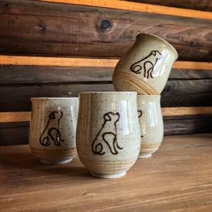 May include: Four speckled ceramic tumblers with a brown dog outline design. The beige cups have a textured surface and a light brown band near the base. The cups are stacked on a wooden surface against a wooden background.