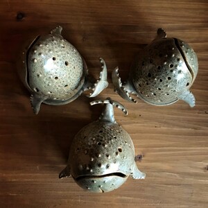 May include: Three ceramic whale-shaped bowls with a speckled gray glaze and small holes throughout. The bowls are arranged in a triangular pattern on a wooden surface.