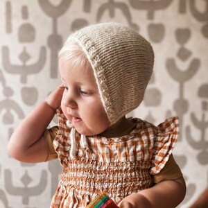 Op de afbeelding: Een baby die een crèmekleurige gebreide muts draagt met een geruit jurkje. De muts heeft een strik die aan de voorkant naar beneden hangt.