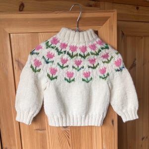 Op de afbeelding: Crèmekleurige trui met een hoge hals en lange mouwen. De halslijn heeft een herhalend patroon van roze tulpen met groene bladeren. De trui hangt aan een metalen hanger tegen een houten achtergrond.