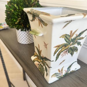 May include: A square white ceramic planter featuring a botanical pattern of green and brown leaves and small flowers. The vessel has a flared rim, a tapered body, and small protruding handles on the sides, showcasing a glossy finish throughout.