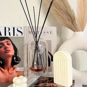 May include: A white, arched candle with a ribbed design sits on a book with a magazine and a glass diffuser with black reeds. The diffuser has a brown liquid inside and the label reads "Bouquet Poudre".