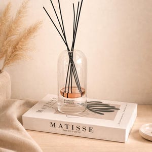 Könnte beinhalten: Ein klarer Glasdiffusor mit schwarzen Stäbchen und einem kupferfarbenen Sockel, der mit "Bouquet Poudre" beschriftet ist. Der Diffusor steht auf einem Buch mit dem Titel "Matisse The Cut-Outs" auf einer Holzoberfläche. Getrocknete Pflanzen sind im Hintergrund.