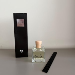 Può includere: Una scatola nera con il testo "No. 3" e la parola "Aura" in un font corsivo. Una bottiglia di vetro trasparente con un tappo di legno e il testo "No. 3 Premium Reed Diffuser" è accanto alla scatola. Bastoncini diffusori di canna neri sono su una superficie grigia.