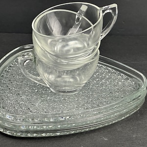 May include: A stack of clear glass teacups with handles and a small spoon inside, resting on a set of matching triangular-shaped glass saucers. The saucers have a textured, patterned surface.