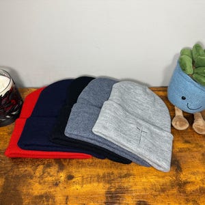 May include: Four beanie hats stacked on top of each other. The top hat is gray, the second hat is blue, the third hat is black, and the bottom hat is red.