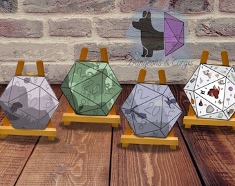 May include: Four D20 dice on small wooden easels. Each die is a different color and design, with numbers and illustrations. The dice are displayed against a brick wall and wooden floor background. The text "Dungeons & Corgis" is in the background.