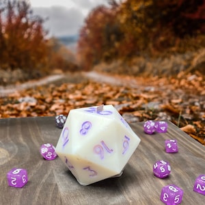Könnte beinhalten: Eine weiße Kerze in Form eines zwanzigseitigen Würfels mit violetten Zahlen auf einer Holzoberfläche mit mehreren kleineren violetten Würfeln.