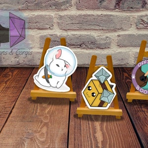 May include: Three small wooden easels each displaying a different sticker. The first sticker is a white rabbit wearing a space helmet and holding a carrot. The second sticker is a yellow cube with a smiling face and dice on top. The third sticker is a brown dog with a purple paw print and the text "OFFICIAL BOOPER".