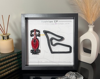 May include: A framed artwork featuring the Austrian Grand Prix. The black frame surrounds a white background with a red race car model and a track layout. Text includes "Austrian GP" and race details. A clear glass car model sits below.