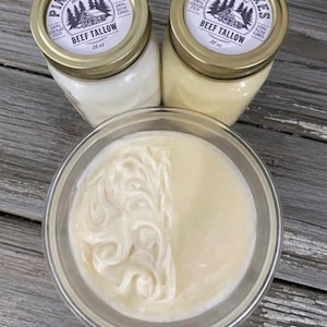 May include: Two glass jars of beef tallow with gold lids and labels that read "Pine Lakes Homestead Beef Tallow 16 oz." A clear glass bowl contains beef tallow, with a swirl design on one side and a smooth texture on the other.