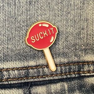 May include: A gold-colored pin shaped like a lollipop with a red candy part that says "SUCK IT" in white letters. The lollipop stick is white with a gold outline.