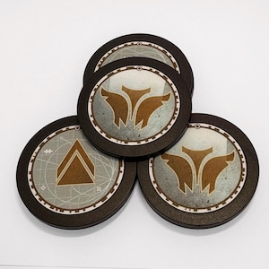 May include: Three circular tokens with dark brown edges and a central metallic silver inlay. Two tokens feature a gold-coloured winged emblem, while the third displays a gold-coloured triangle icon. Each token has a thin outer ring with markings.
