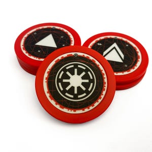 May include: Several red, circular game tokens with black and white designs. One token shows a white triangle, and another has a black and white Imperial symbol. These tokens are likely for a board game.