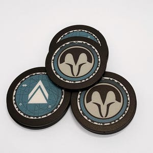 May include: Three circular coasters with black edges and a central design. Two coasters feature a stylised tan ram head icon on a blue background, while the third displays a white triangle symbol over a blue map-like pattern with grid lines.